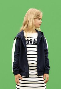 Giacca zip-up blu navy con maniche bianche e accento verde; indossata sopra una maglietta a maniche lunghe bianca e a righe. Sfondo verde solido.