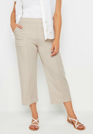 Kvinde iført beige cropped bukser med vide ben og lommer, hvid kortærmet bluse med øjeletdetaljer og hvide flade sandaler med stropper.