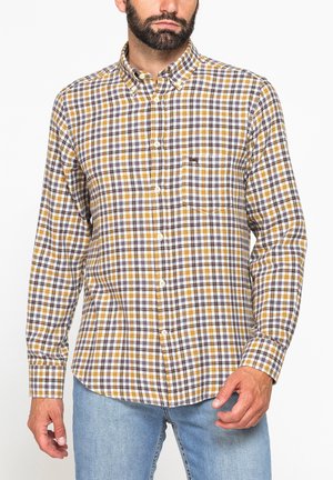 Uomo che indossa una camicia a quadri gialla, marrone e bianca con maniche lunghe e jeans azzurri chiari, in piedi davanti a uno sfondo bianco.