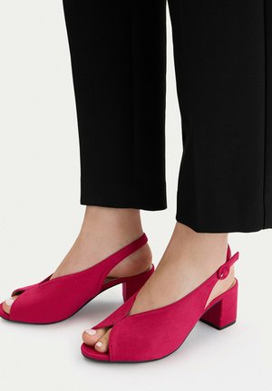 Pies con pantalones negros que llevan zapatos de tacón bloque abierto en rosa brillante sobre un fondo claro.