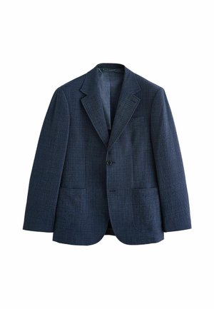 Junger Mann mit kurzen dunklen Haaren, der einen dunkelblauen karierten Blazer über einem bordeauxfarbenen, geknöpften Hemd vor einem einfachen hellen Hintergrund trägt.