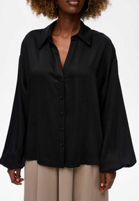 Zwarte overhem shirt met een kraag, lange, luchtige mouwen en een gladde, matte textuur. Voorzien van een knoopsluiting aan de voorkant.