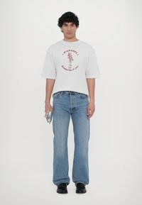 Hvit oversized t-skjorte med korte ermer og rød grafisk tekst, sammen med lys blå denimjeans, komplett med svarte sko og holder en liten sølvgjenstand.