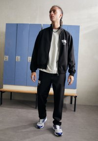 Svart jakke med glidelås og hvit logo, kombinert med matchende bukser. Bruker en lys grå t-skjorte og grå sneakers med blå aksenter.
