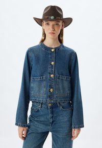 Chaqueta de mezclilla con mangas largas, dise�ño corto, dos bolsillos frontales, botones dorados y un color azul desgastado, combinada con pantalones de mezclilla y un sombrero marrón.