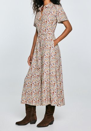 Mujer con vestido floral de manga corta con botones en el frente, cinturón en la cintura y botas marrones de ante hasta media pantorrilla, de pie con las manos en los bolsillos.