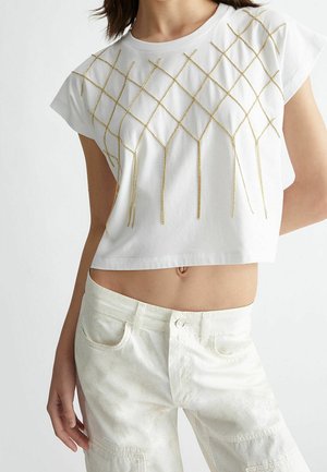 Femme portant un t-shirt court blanc avec un motif de franges en chaîne dorée et un pantalon blanc taille haute, debout avec un bras détendu.