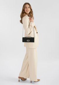 Bolso de mano negro con asa de cadena, textura suave, diseño rectangular y herrajes plateados, exhibido junto a un traje beige y tacones marrones.