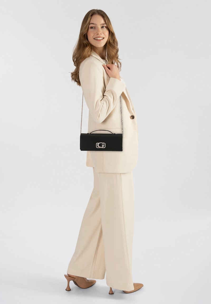 Bolso de mano negro con asa de cadena, textura suave, diseño rectangular y herrajes plateados, exhibido junto a un traje beige y tacones marrones.