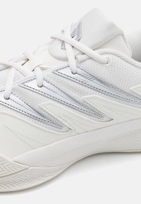 Chaussure de sport blanche avec une tige texturée aux accents argentés, une semelle à motifs, un design épuré avec des lacets visibles et un logo.