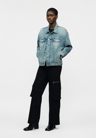 Γυναίκα όρθια με τα χέρια στις τσέπες, φορώντας ανοιχτό μπλε τζιν μπουφάν, μαύρο παντελόνι cargo με φερμουάρ και μαύρα μποτάκια με τακούνι.