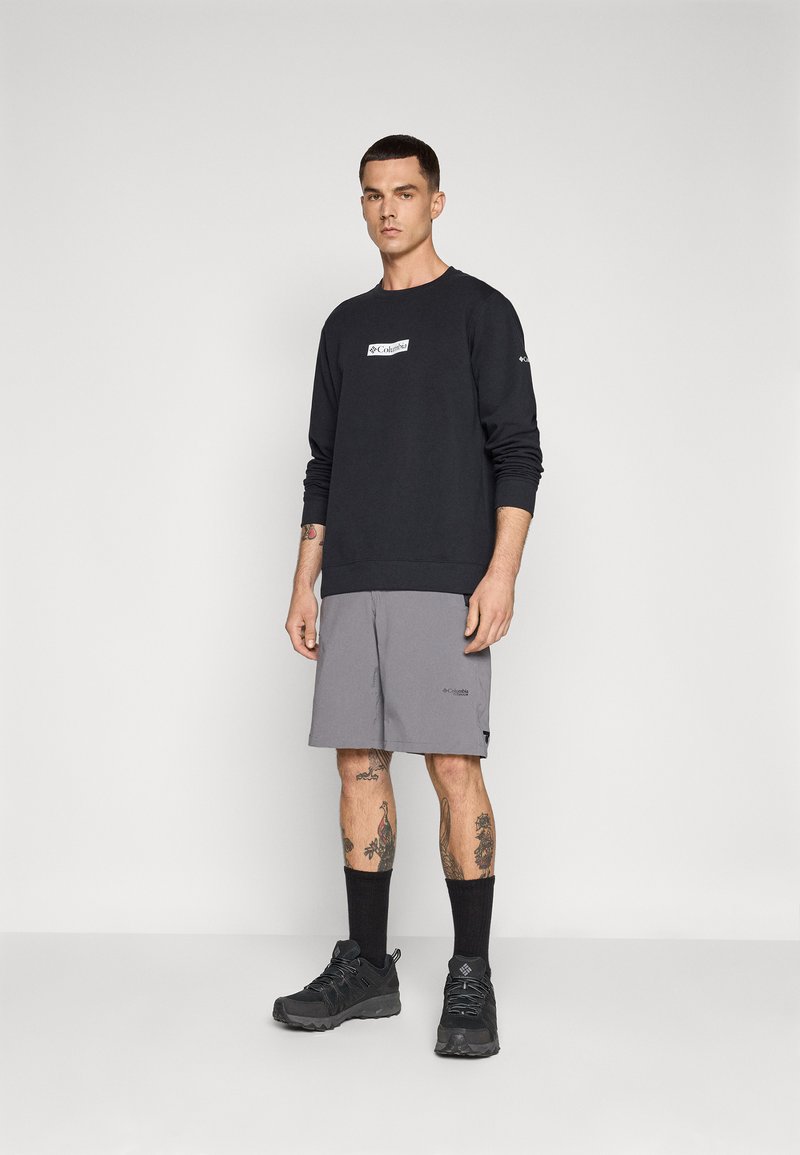 Sweat-shirt noir avec logo, shorts gris, baskets noires et chaussettes longues. Tatouages visibles sur les bras et les jambes. Debout sur un fond uni.