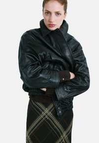 Chaqueta de cuero negra con cuello alto, con elástico en el dobladillo. Combinada con una falda de cuadros en colores verde, marrón y beige.