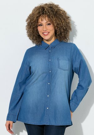 Lächelnde Frau mit lockigem Haar trägt ein langärmeliges blaues Jeanshemd mit Knöpfen und Brusttasche und steht vor einem schlichten weißen Hintergrund.