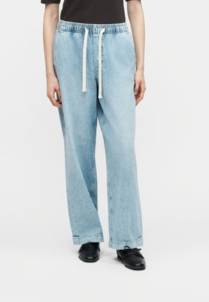 Person trägt hellblaue Jeans mit weitem Bein, elastischem Bund und weißem Kordelzug, kombiniert mit schwarzen Schnürschuhen und schwarzem Oberteil.