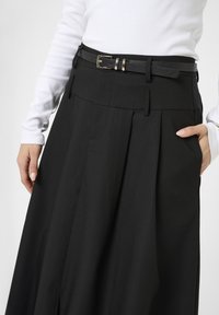 Falda negra con pliegues que cuenta con bolsillos laterales, una cinturilla con pasadores para cinturón y una textura suave. Combinada con una blusa ajustada de manga larga blanca.