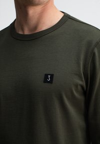 Olivengrünes Langarm-T-Shirt aus weichem Stoff mit Rundhalsausschnitt, versehen mit einem kleinen schwarzen Quadrattuch mit einem weißen 'J'-Logo.