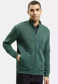 Zielona kurtka zapinana na zamek z wysokim kołnierzem, prążkowanymi mankietami i dwoma bocznymi kieszeniami. Gładki materiał o luźnym kroju. Nosi się ją na ciemnej koszuli.