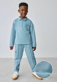 Himmelblaues Zip-Fleece-Sweatshirt mit einer Tasche und passenden Cargo-Hosen, die aus strukturiertem Stoff bestehen, sowie weißen Sneakern mit einer Gummisohle.