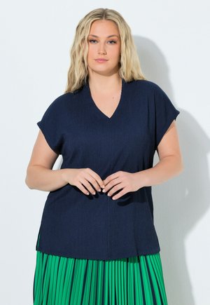 Blondynka o falowanych włosach ubrana w granatowy krótki rękaw z dekoltem w serek i zieloną plisowaną spódnicę, stoi na tle białej ściany.