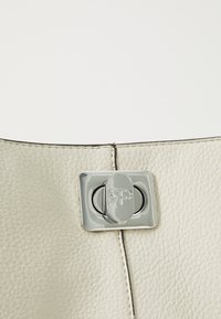 Kremowa skórzana torebka o teksturowanej powierzchni, z zamkiem z srebrnego metalu oraz szczegółami wytłoczonymi z logo. Czysty, minimalistyczny design.