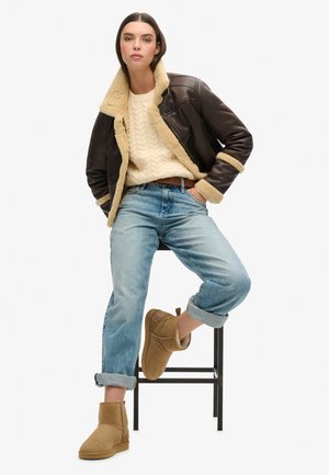 Femme assise sur un tabouret noir, portant une veste en peau retournée marron foncé, un pull crème à torsades, un jean bleu retroussé et des bottines camel.