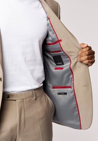 Beige Sakk mit roten Akzenten und grauem Futter, ausgestattet mit mehreren Innentaschen. Kombiniert mit einem weißen T-Shirt und beigen Hosen.