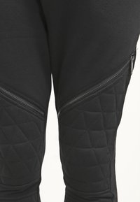 Černé tepláky s prošívanou kolenní panelem, mají boční zipy a hladkou texturu látky. Přiléhavý střih s úzkou nohavicí.