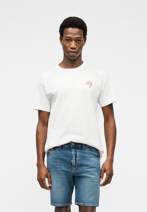 Fiatal férfi fehér pólóban, kis logóval és kék farmer rövidnadrágban, egyszínű világos háttér előtt áll.