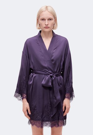 Femme portant un peignoir en satin violet longueur genou avec bordure en dentelle aux manches et à l'ourlet, noué à la taille avec un nœud.