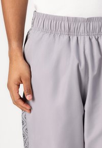Lichtgrijze sportbroek van glad, lichtgewicht materiaal met een elastische tailleband en een zijpaneel met een herhaald logo patroon.