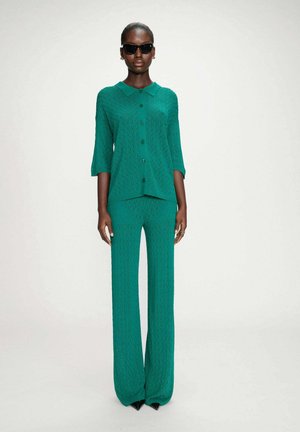 Femme noire portant un pantalon large texturé bleu sarcelle et un haut boutonné assorti, accessoirisée avec des lunettes de soleil noires et des boucles d'oreilles, debout devant un fond blanc.