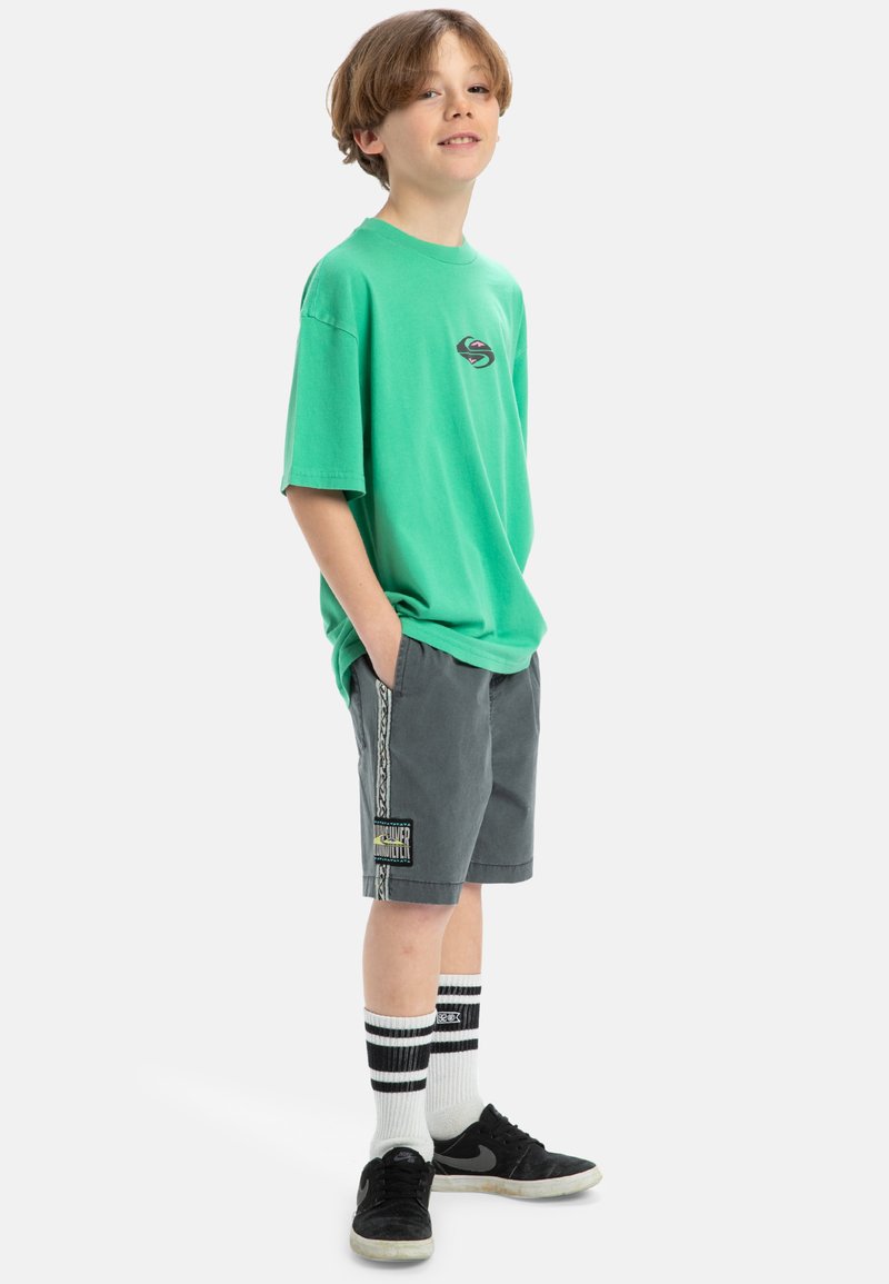 Zelené oversized tričko z bavlny, šedé kraťasy s bočním pruhem a logem, černé tenisky a bílé ponožky s viditelnými černými pruhy.