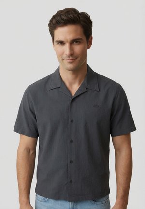 Homme aux cheveux foncés portant une chemise boutonnée sombre à manches courtes avec col et un jean bleu clair sur un fond clair uni.