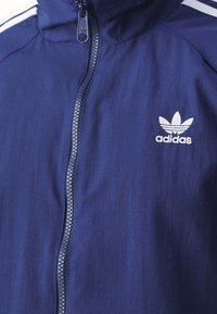 Navy blue Adidas jacket featuring a high collar, front zipper, and white stripes along the shoulders; logo displayed on the left chest.