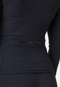 Svart ribbad träningsöverdel med en åtsittande silhuett, långärmar och en dragkedjeficka på baksidan för förvaring.