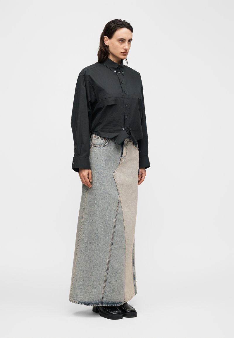 Femme portant une chemise noire boutonnée rentrée dans une jupe longue en denim bicolore, avec des chaussures noires à bout carré, debout devant un fond uni.