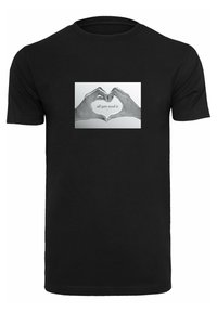 Svart bomull t-skjorte med et grafisk motiv av to hender som danner et hjertesymbol, med teksten "alt du trenger er..." i en lys skrifttype.
