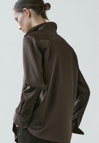 Camicia a maniche lunghe a righe marroni con collo alto e polsini abbottonati, realizzata in tessuto liscio; presenta una vestibilità comoda e un orlo asimmetrico.