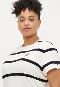 Fehér csíkos póló, fekete vízszintes vonalakkal, kerek nyakkivágással, rövid ujjal, apró logóval a mellrészen és kiemelő részletekkel.