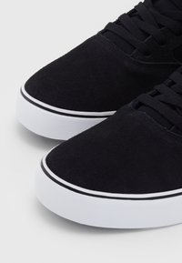 Baskets en daim noir avec semelles en caoutchouc blanc et fermetures à lacets noires, vues de face en angle.