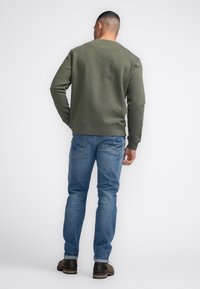 Olijfgroen sweatshirt met geribbelde manchetten en zoom, gecombineerd met lichtblauwe jeans en bruine laarzen, gezien van achteren tegen een effen achtergrond.
