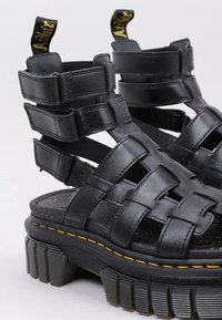 Sandalias de plataforma negras con amplias correas trenzadas, plantilla texturizada, suela de goma gruesa y detalle de costura amarilla. Cuenta con una pestaña en la parte posterior.