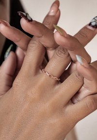 Różowo-złoty pierścionek z kręconym bandem na dłoni z misternie wypielęgnowanymi paznokciami w różnych kolorach i wzorach. Widoczna jest teksturowana skóra.