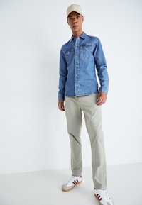 Camicia di denim blu chiaro, con due tasche sul petto e chiusura a bottoni. Abbinata a pantaloni beige e sneakers bianche con strisce rosse.