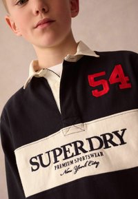 Černé a krémové ragbyové tričko s bílým vyšitým logem "SUPERDRY" a červeným číslem "54" na prsou; bavlněný materiál a límečkový design.