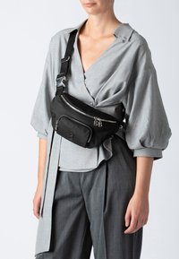 Borsa a tracolla nera in nylon con tasche anteriori con zip, logo in rilievo e tracolla regolabile. Texture liscia, design compatto, indossata sopra una maglietta grigia.