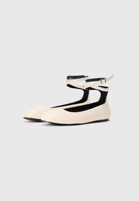 Une paire de ballerines plates beige avec des brides de cheville et de petites boucles, présentées sur un fond gris clair uni.