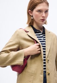 Νεαρή γυναίκα που φορεί μπεζ σακάκι και ριγέ πουκάμισο, κρατώντας μικρή κόκκινη τσάντα ώμου, κοιτάζει μπροστά με ουδέτερη έκφραση.