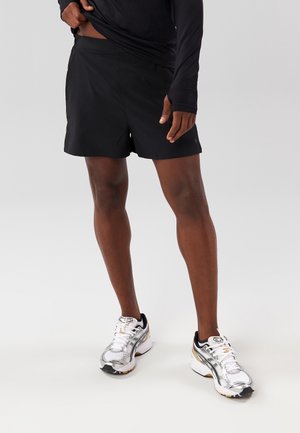 Mann trägt schwarze Sportshorts, schwarzes langärmliges Oberteil und weiße Laufschuhe und steht vor einem neutralen Hintergrund.